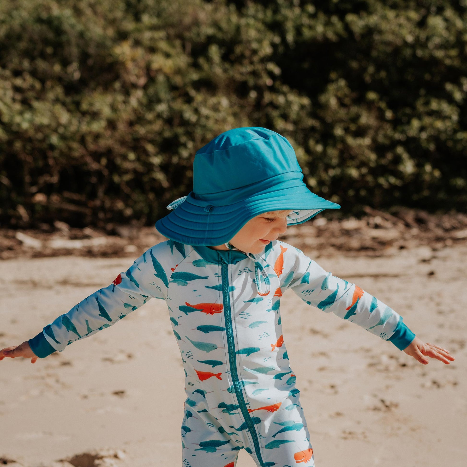 Toddler wearing Pipi Swimwear narwhal full length sunsuit and matching wide brim aqua reversible swim hat